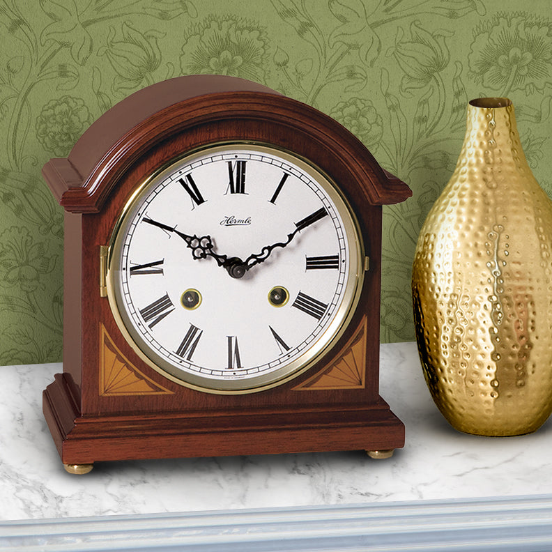 Wooden barrister style mantel clock with white face and gold accents on a marble surface with a gold vase and green floral wallpaper.