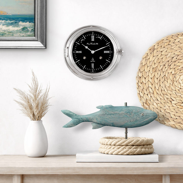Nickel and black Ship's Bell clock on a wall above a table with a fish sculpture and nautical rope.