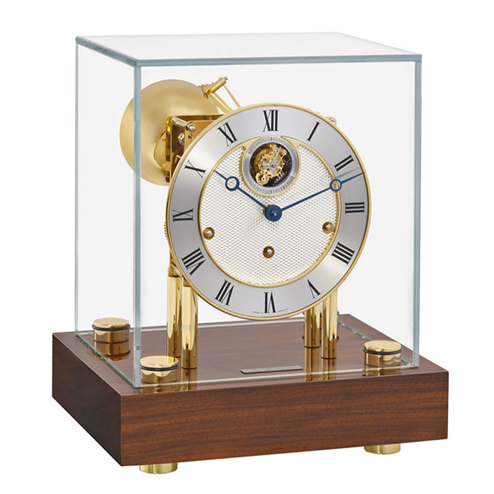 A luxury desk clock with a polished brass body, tourbillon, metallic dial with Roman numerals, and a walnut wood base, all enclosed in a clear glass display case.