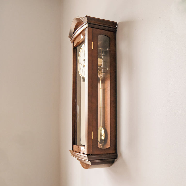 Side view of wooden wall clock with glass panes and brass pendulum hanging on white wall