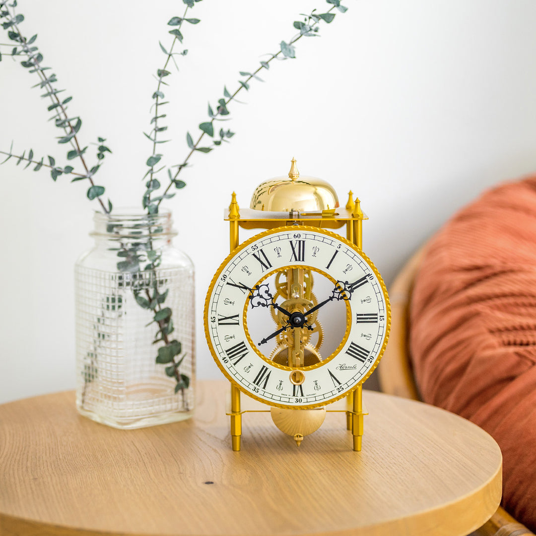 Brass clock sitting on wood table with green in jar plant and soft orange chair in background 