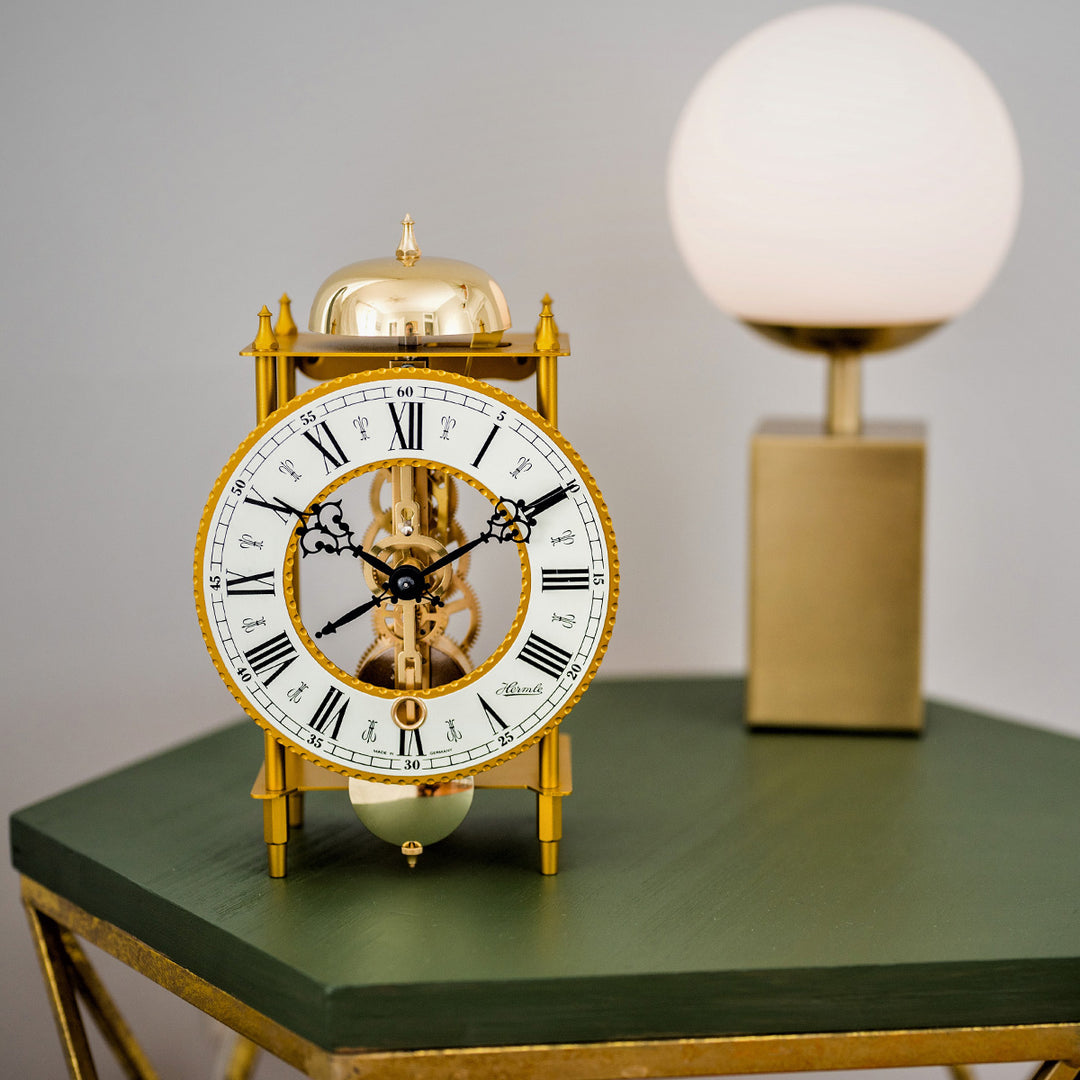 Brass mantel clock with roman numeral face on green table in front of lamp