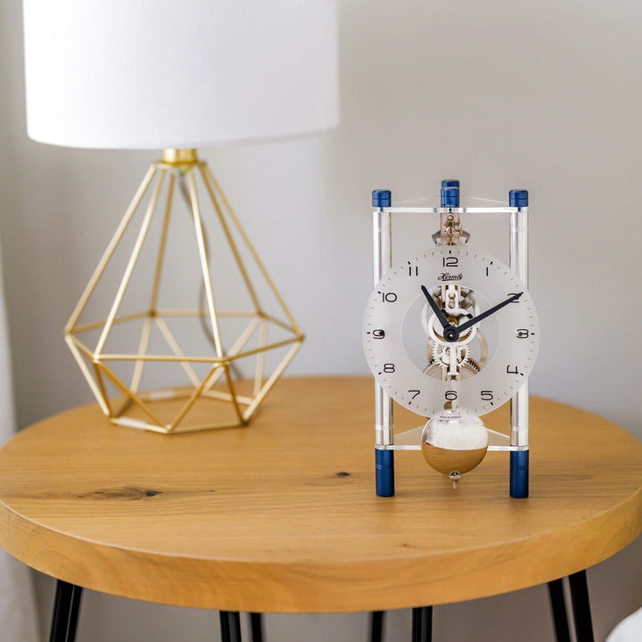 Silver and blue mechanical clock with intricate gears visible on wood table next to gold lamp