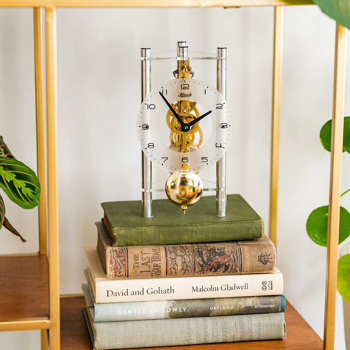 Silver and brass mechanical clock with glass face and intricate gears visible on stack of books