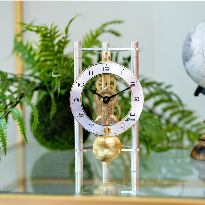 Silver and brass mechanical clock with intricate gears visible sitting on glass table in front of green plants