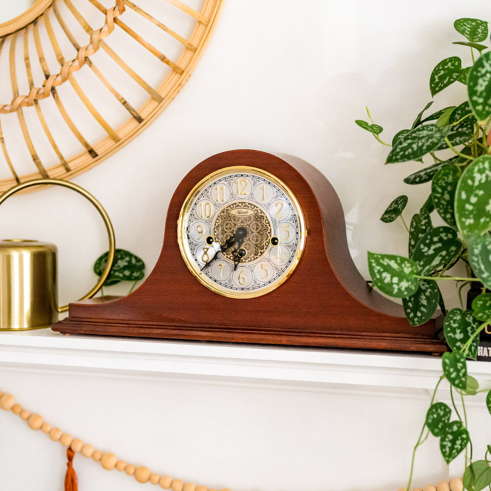 Cherry wood mantel clock with ornate hands on white mantel next to green plants. 