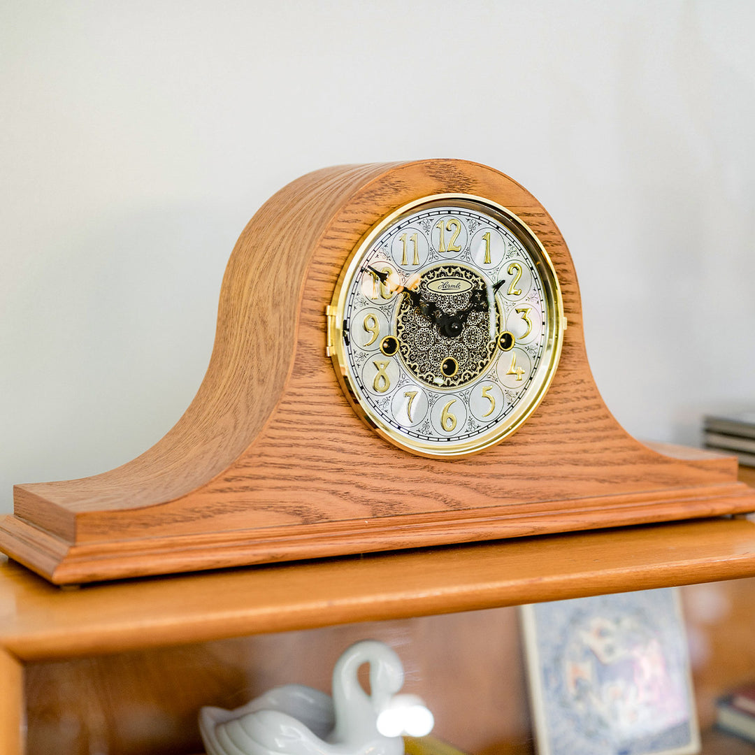 Light Oak mantel clock with brass highlights on wooden dresser in front of white wall. 