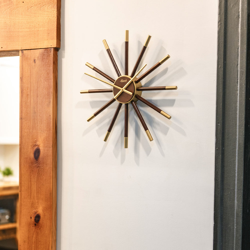 Sunburst style clock with brass highlights hanging on white wall next to wooden doorway.