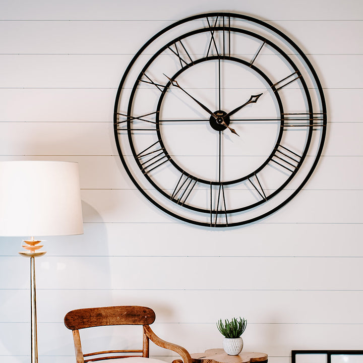 Metal decorative wall clock with Roman numerals on a white wall above chair and lamp