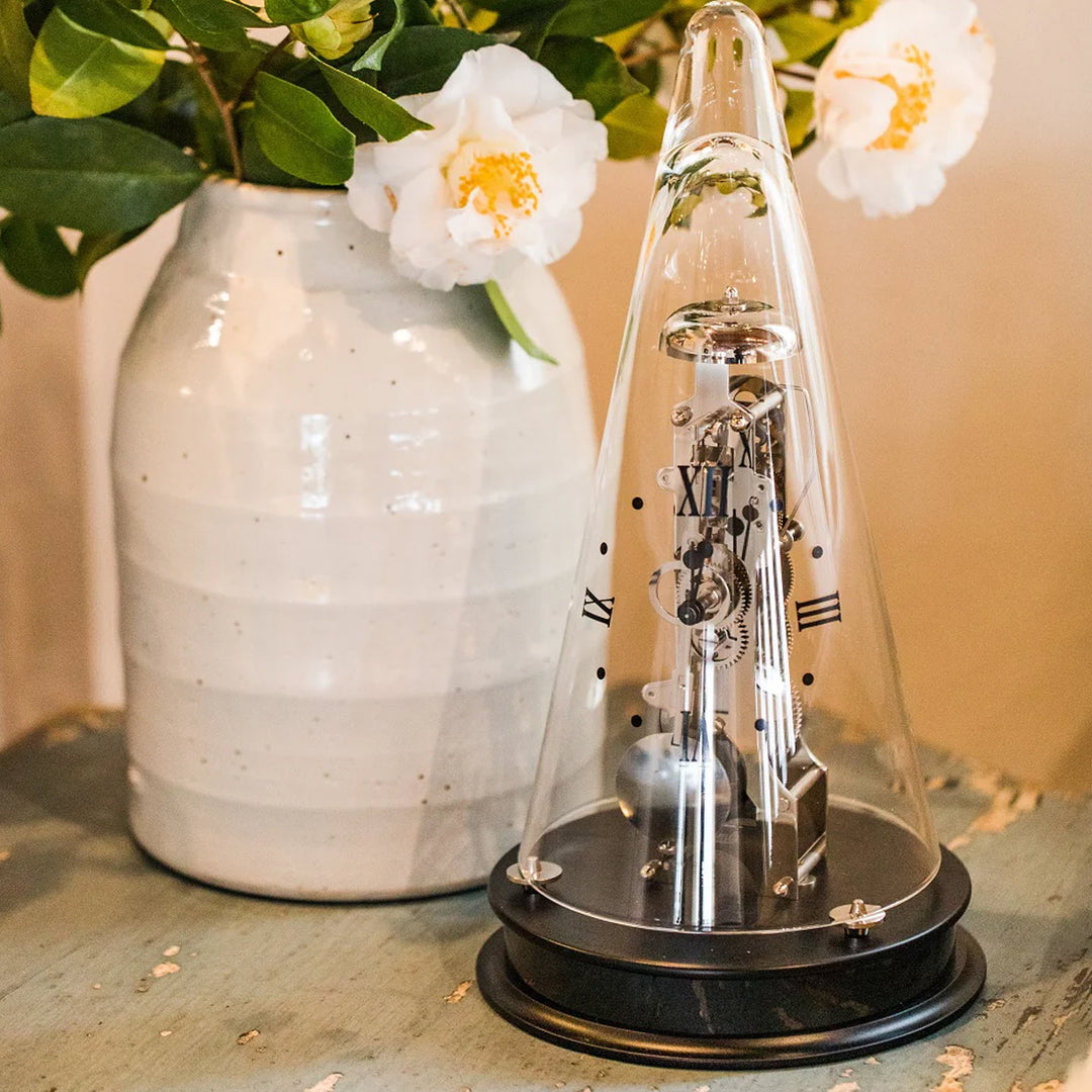 Decorative clock under a glass dome on a table with a vase and flowers in the background