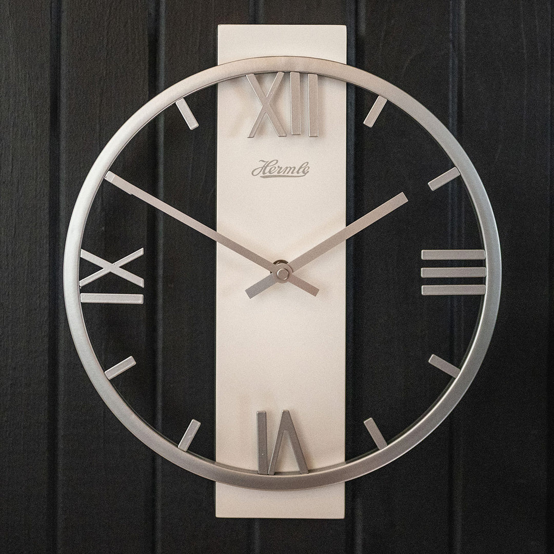 Metal clock with roman numerals, Hermle logo and open frame design hanging on black plank wall.