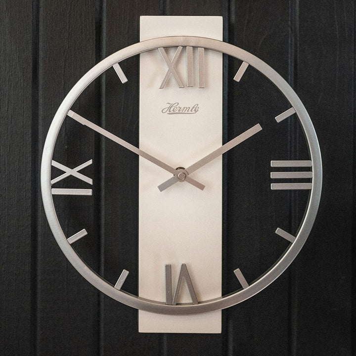 Metal clock with roman numerals, Hermle logo and open frame design hanging on black plank wall.