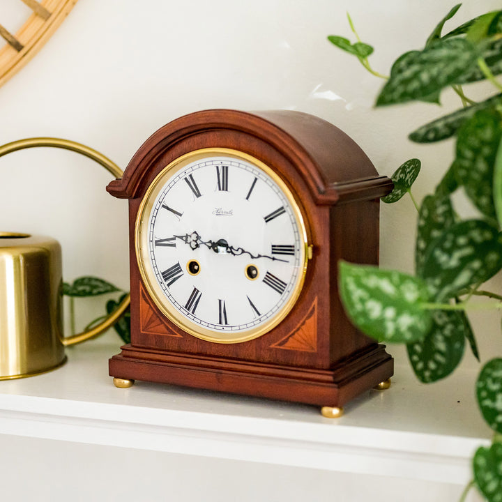 Wooden mantel clock on white shelf with green plant and brass watering can alongside.