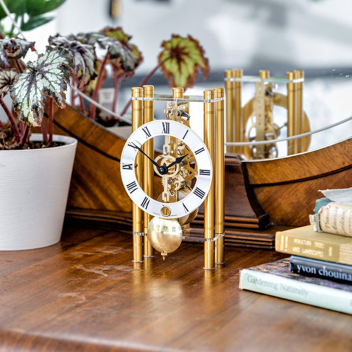 Gold mechanical clock with intricate gears sitting on wooden dresser next to books and potted plant