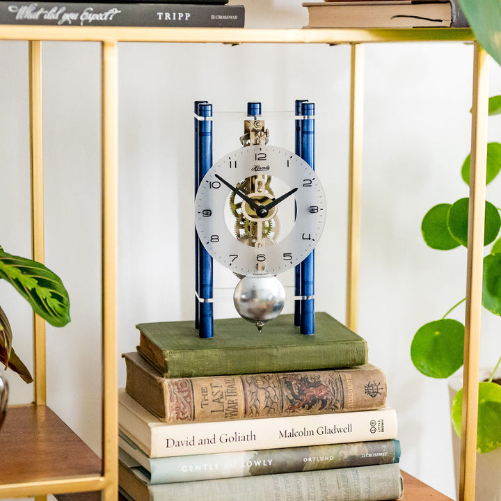 Blue and silver mechanical clock sitting on stack of books next to potted plants 