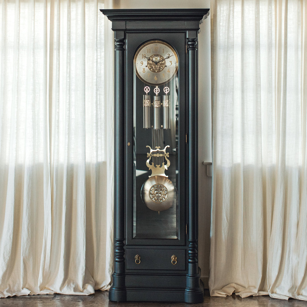 Black grandfather clock with brass and nickel accents against a white curtain background