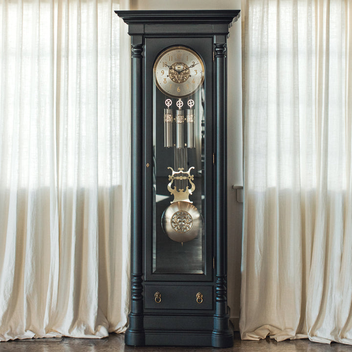 Black grandfather clock with brass and nickel accents against a white curtain background