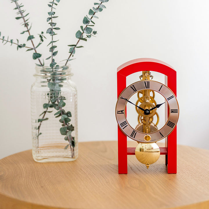 Small red clock with brass mechanism on a wooden surface next to a clear vase with small plant