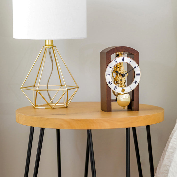 Walnut clock with brass gears and silver face on wooden side table with a geometric lamp 