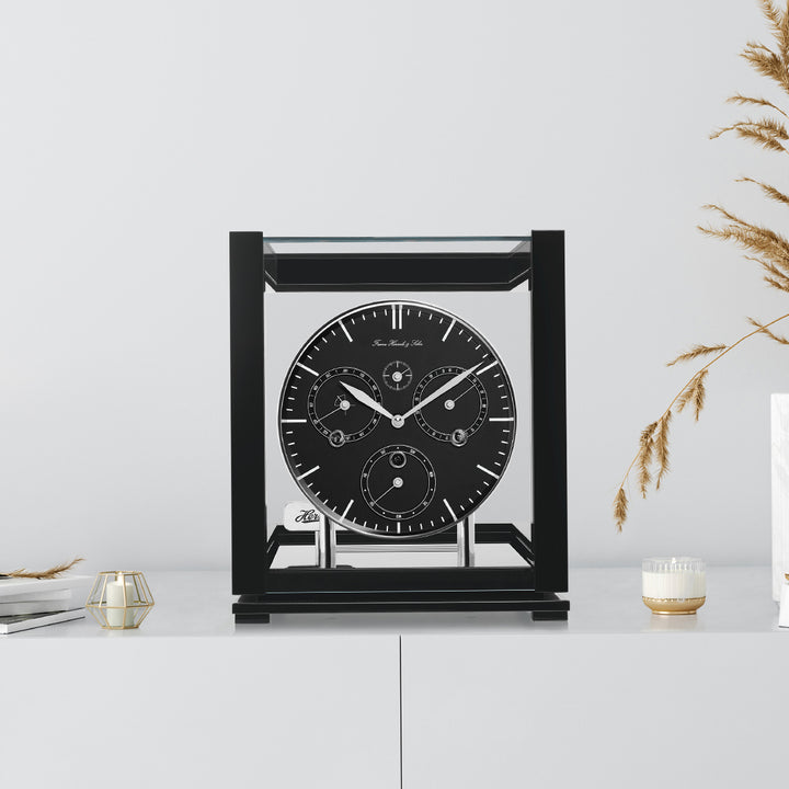 Black perpetual calendar clock with black face on white cabinet with books, candles and tan plants on the side.