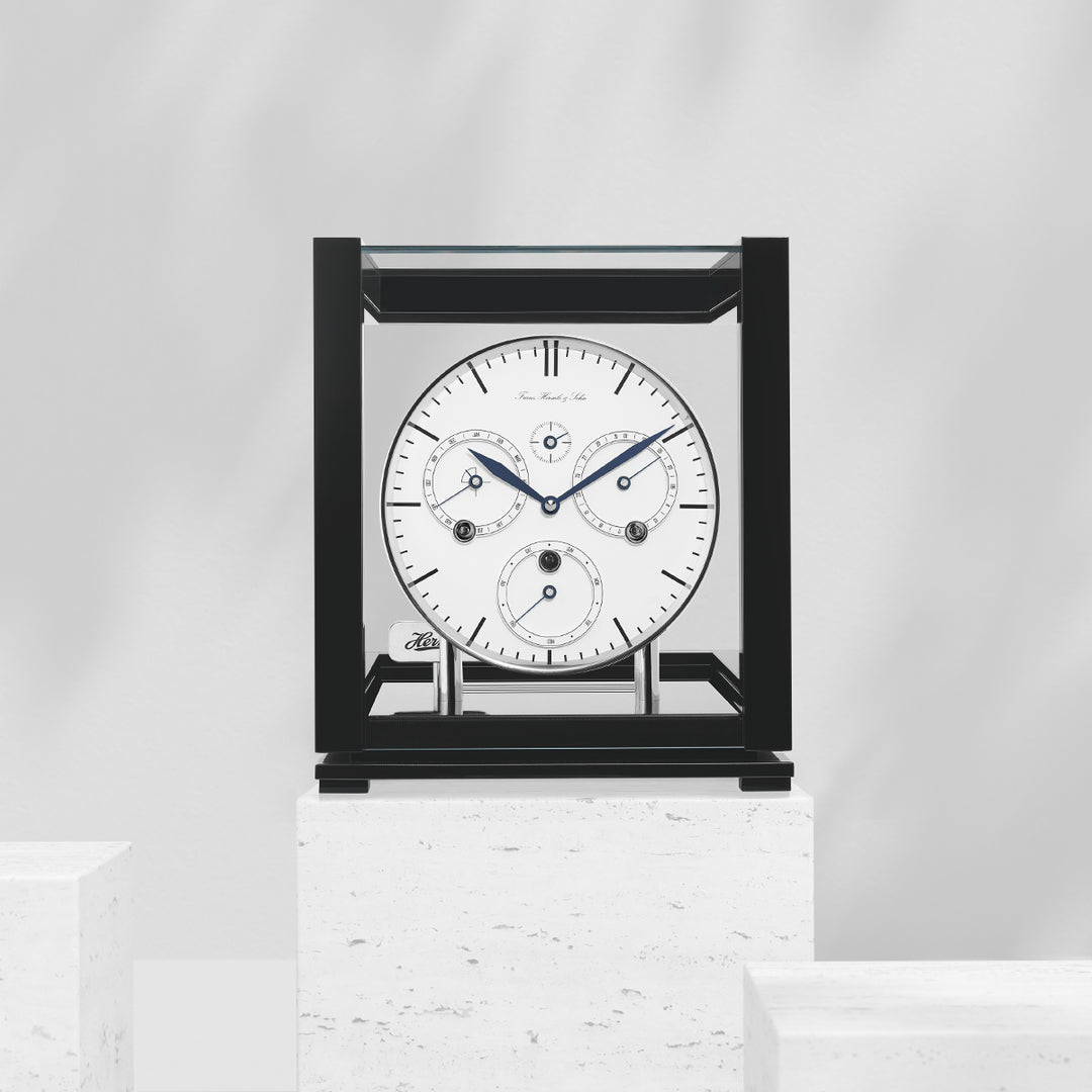 Perpetual calendar clock with white face on white marble pedestal in white room.