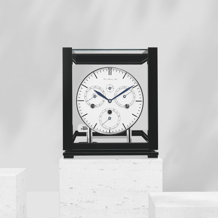 Perpetual calendar clock with white face on white marble pedestal in white room.