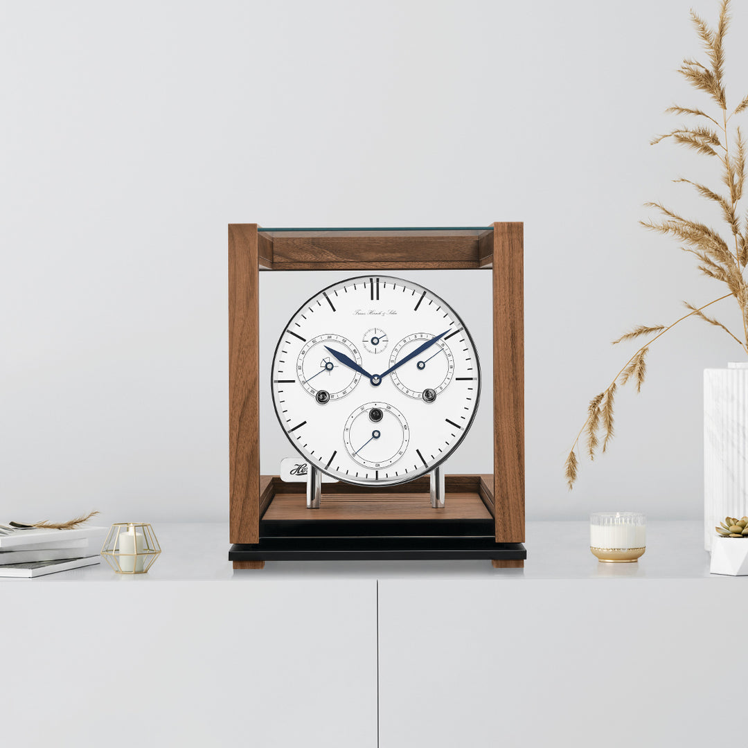 Walnut perpetual calendar clock with white face on white cabinet with books, candles and tan plants on the side.