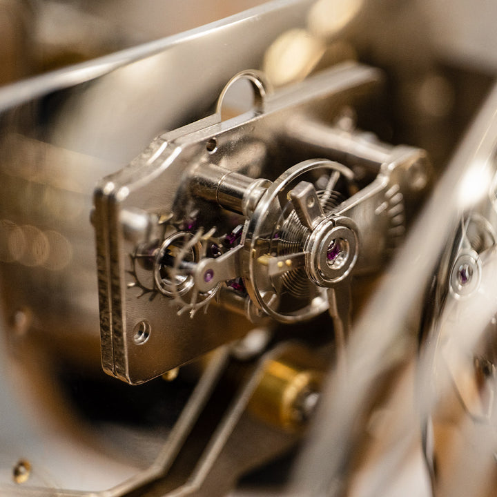 Up close of tourbillion with nickel finish with gears and jewels mounted inside mechanical clock.