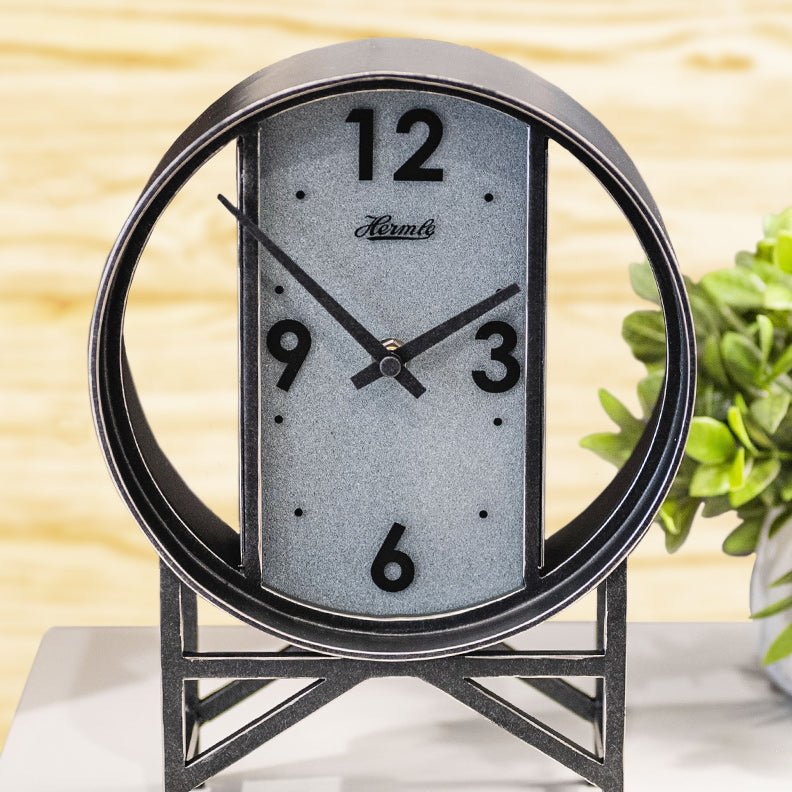 A wrought iron modern desk clock on a white marble desk next to a potted plant.