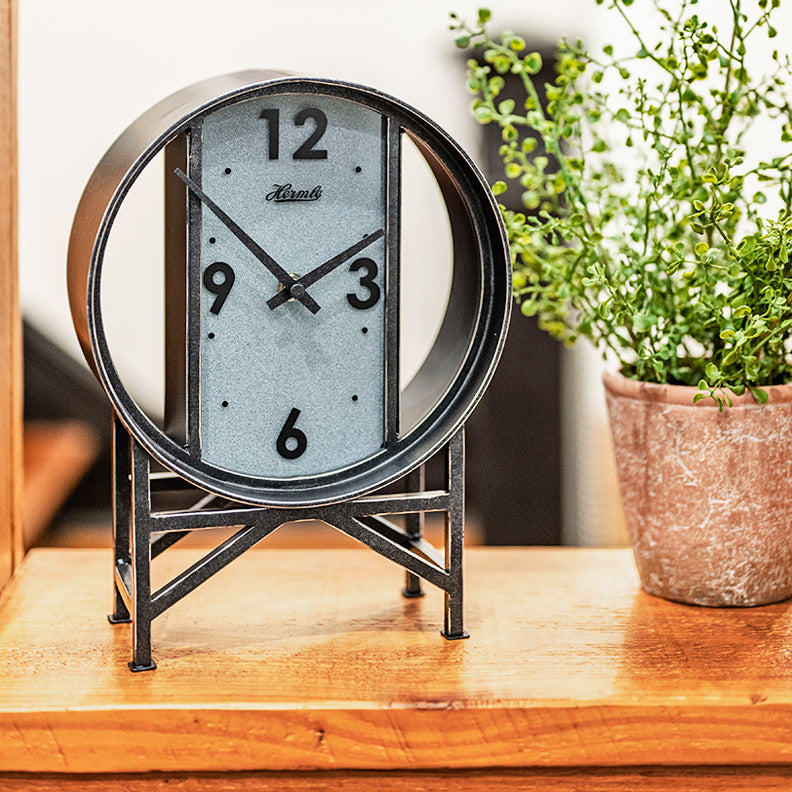A wrought iron modern desk clock on a wooden countertop next to a rustic potted plant.