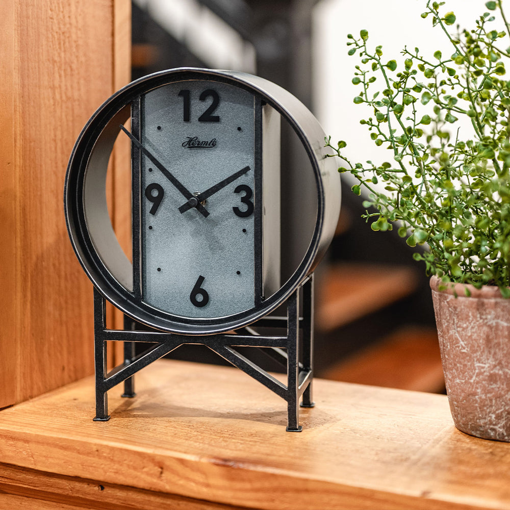 Black metal clock on wooden shelf next to green plant. 