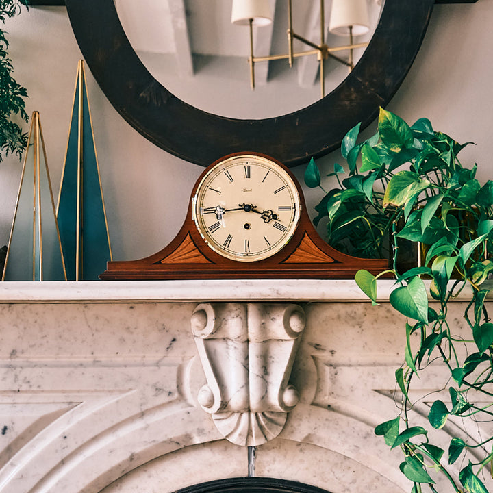 Vintage style clock on marble mantel next to green plant and decorations.