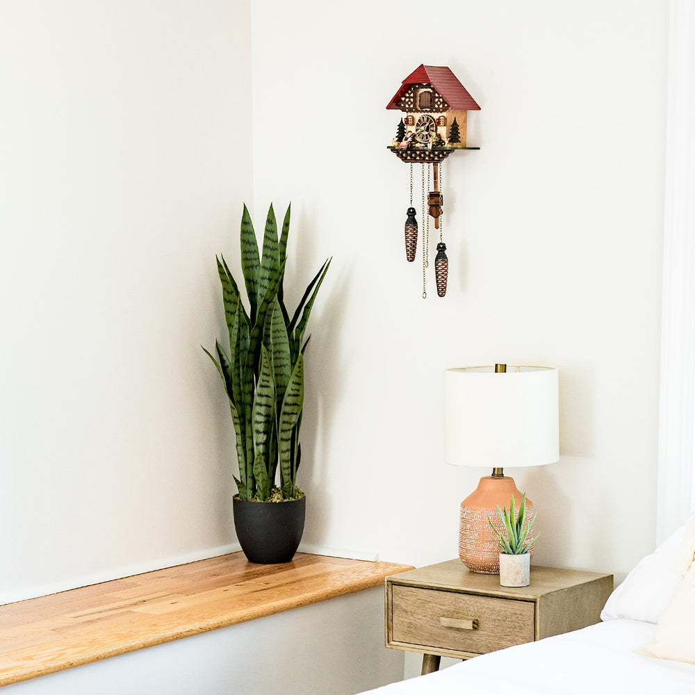 Cuckoo clock with red roof and decorative design hanging on white wall above plant and lamp 