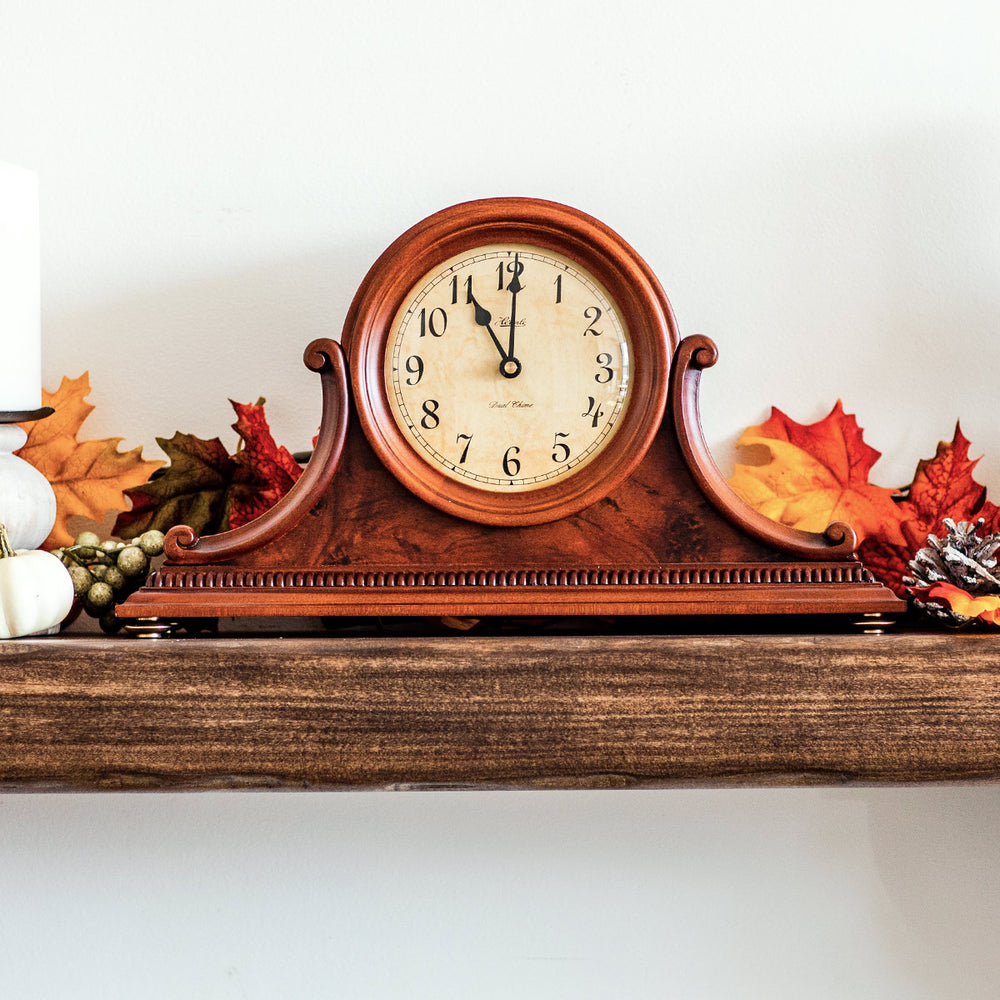 Cherry wood mantel clock on wooden shelf with Hermle logo surrounded by fall leaves on white wall.