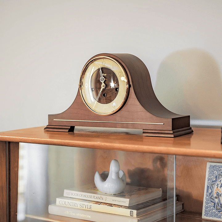 Wooden mantel clock on a wooden shelf with books and decorative decor.