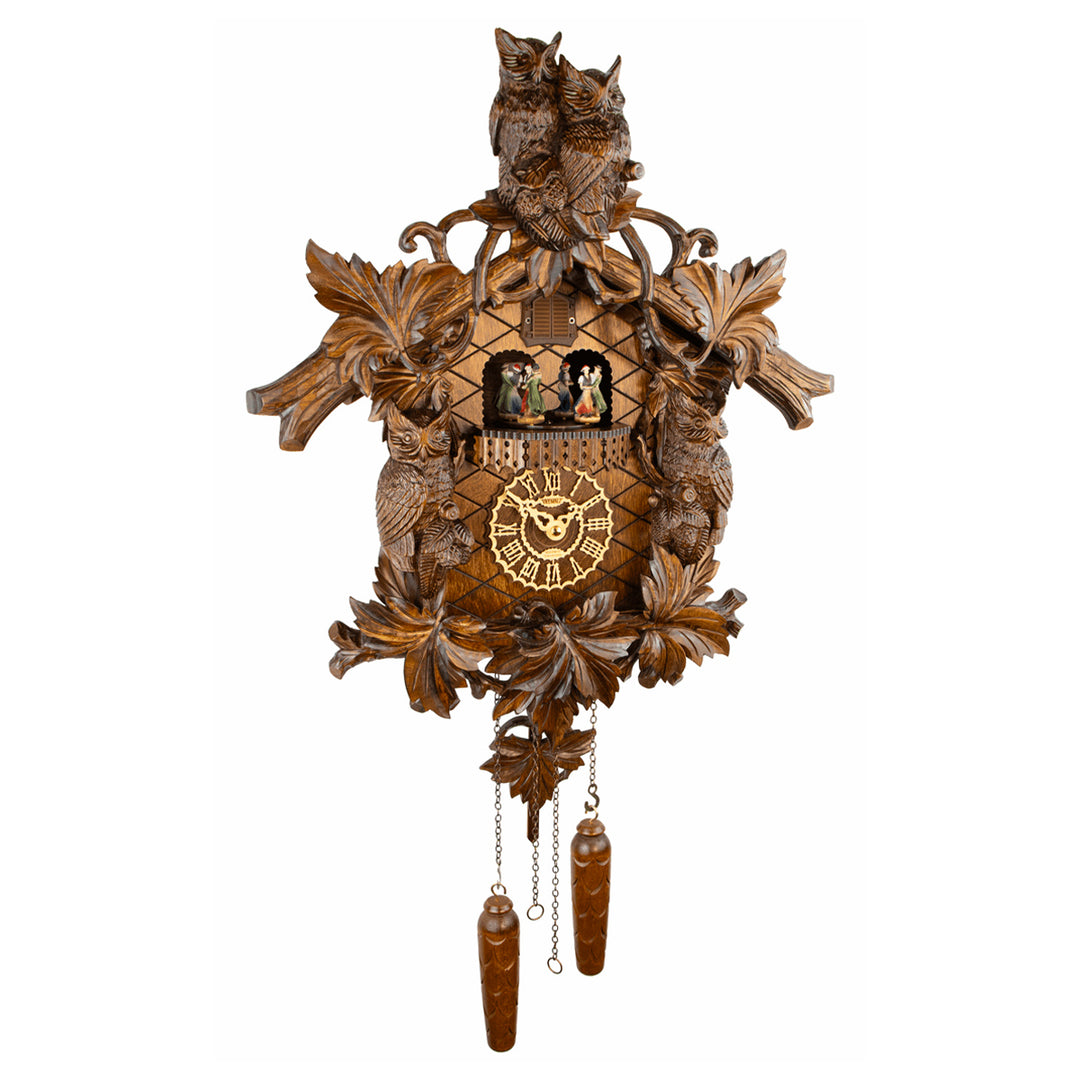 An ornate hand-carved wooden cuckoo clock featuring two owls at the top, leaf and bird motifs, a central clock face with Roman numerals, small figurines in the window, and two dangling wooden weights below, shown against a plain white background.