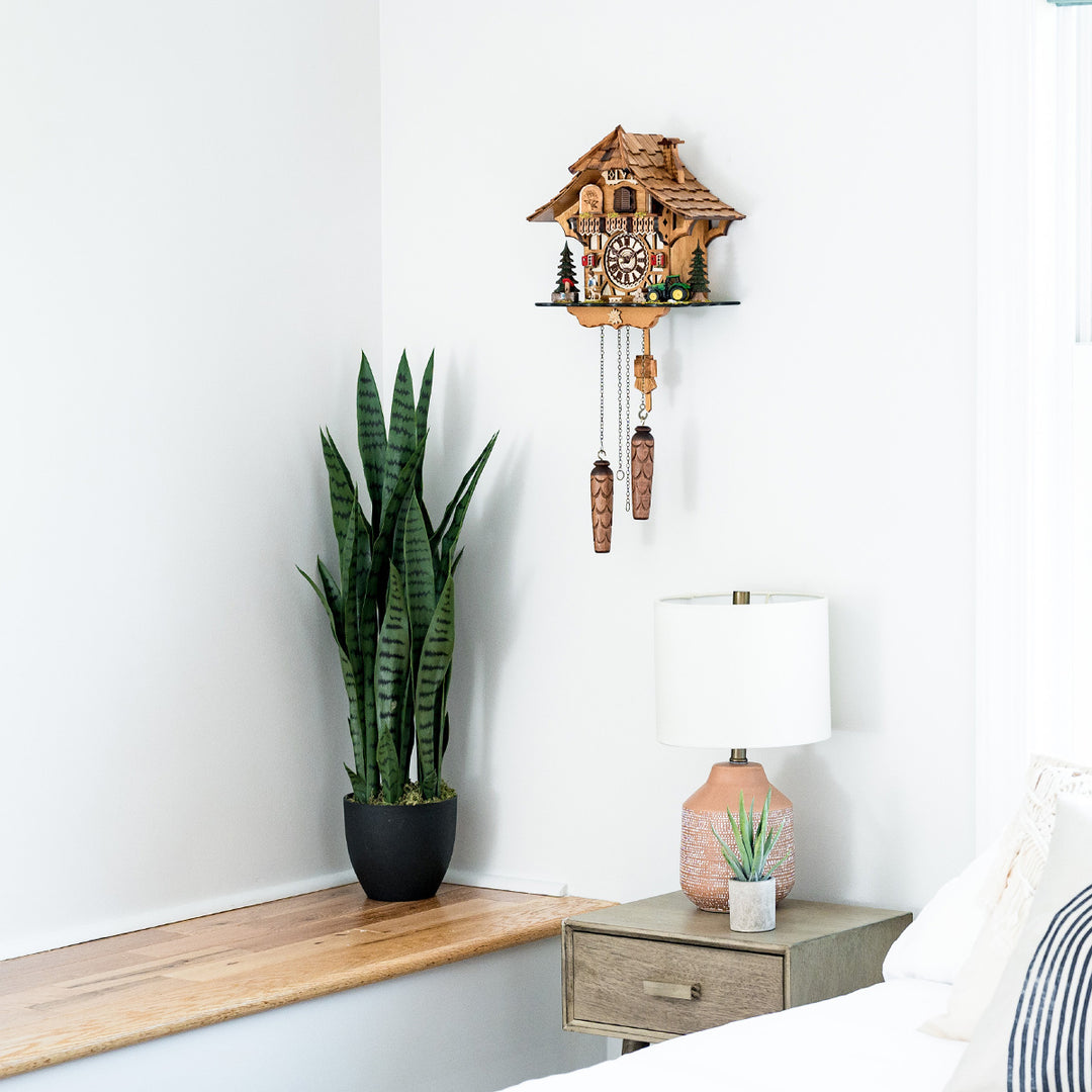 Wooden cuckoo clock on a white wall above a wooden shelf with a potted plant and lamp.