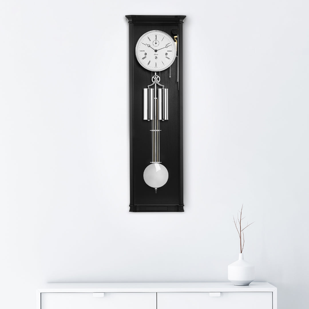 Black regulator wall clock with silver highlights hanging on white wall above white dresser