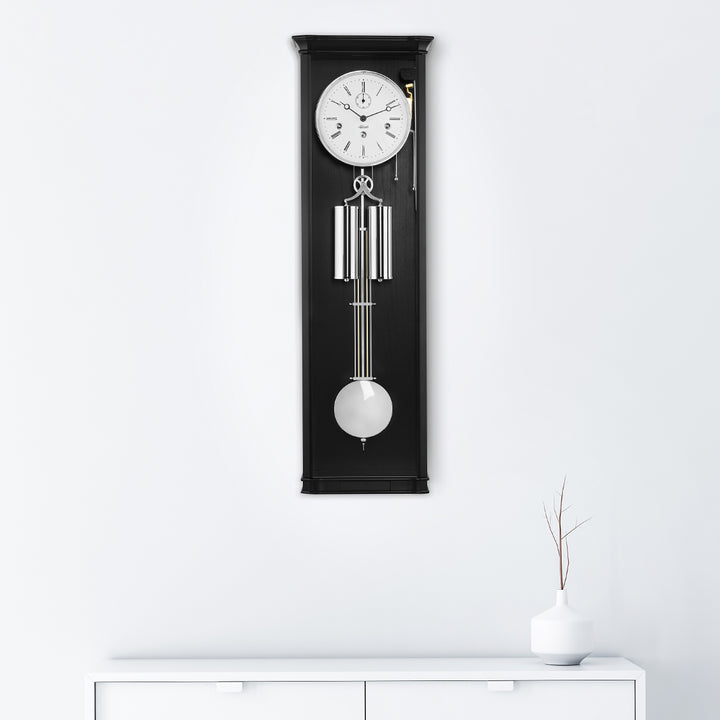 Black regulator wall clock with silver highlights hanging on white wall above white dresser