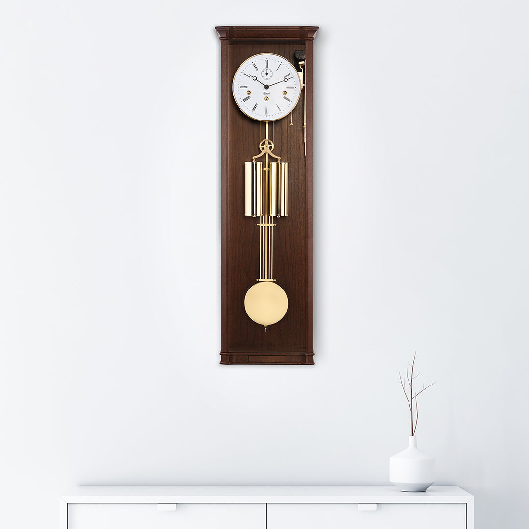 Walnut wood regulator wall clock with brass highlights hanging on white wall above white dresser