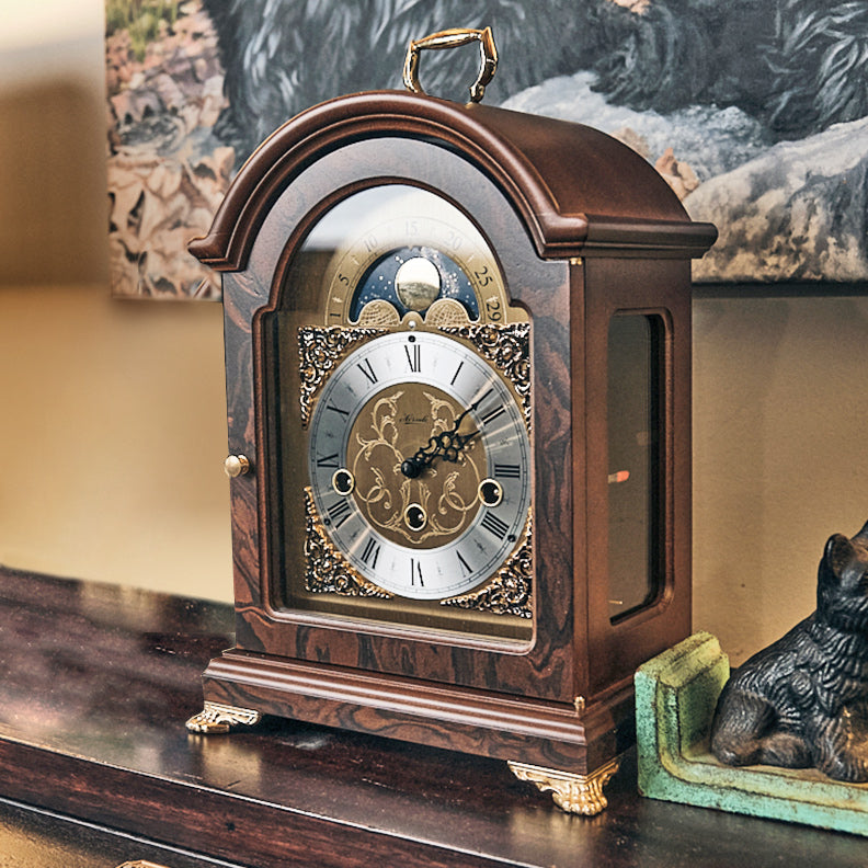 Front, side view of vintage style luxury walnut mantel clock on wood mantel with painting behind it on the wall.