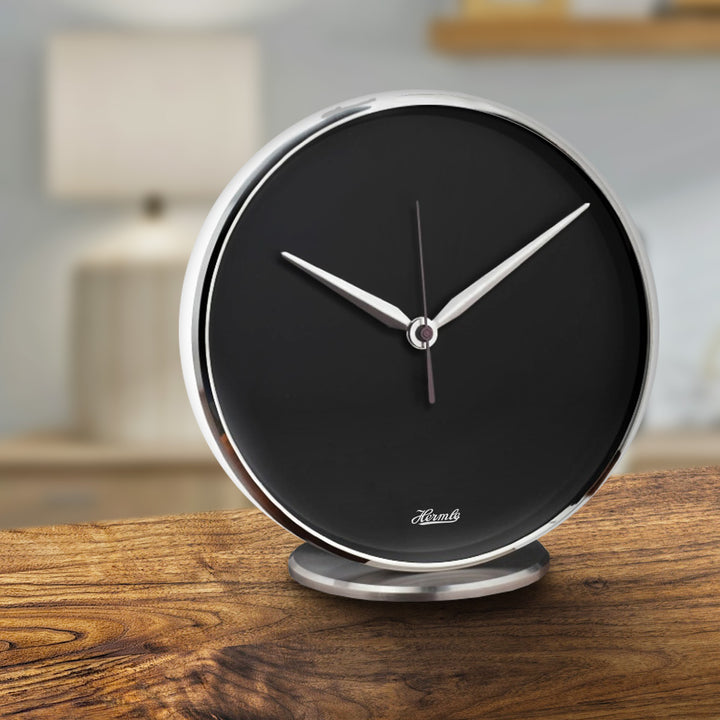 Round desk clock with black dial and metallic rim on wooden table with lamp in background. 